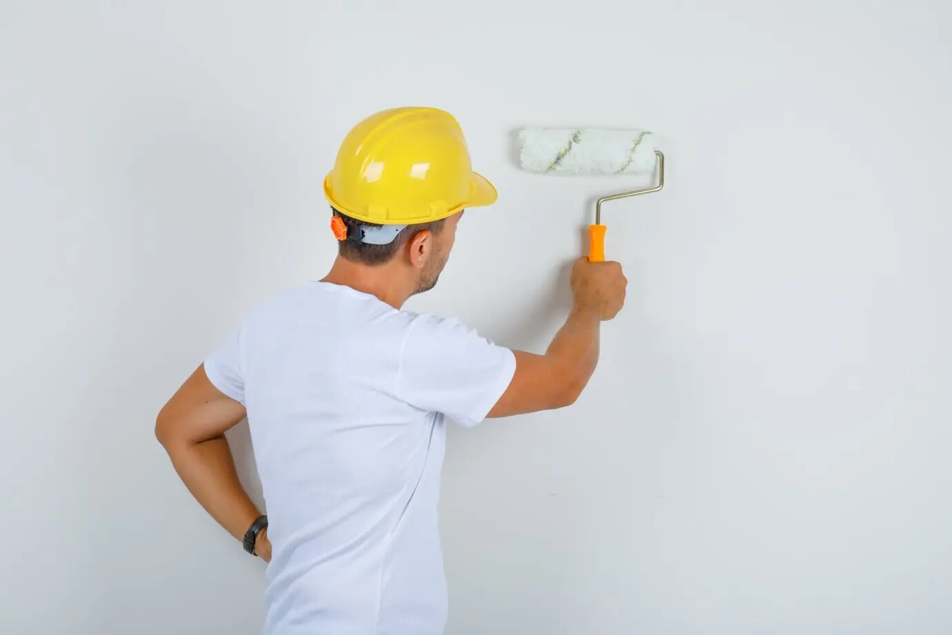 Back view of a male builder wearing a white T-shirt and helmet, painting a wall with a roller and looking busy.